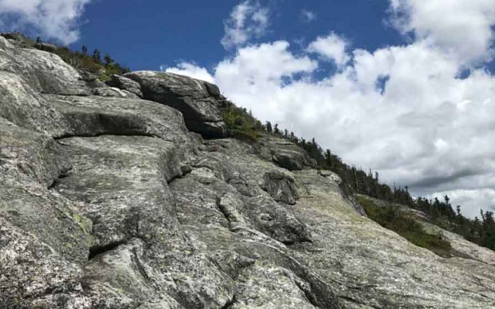a gray rock wall juts up toward a blue sky with some white fluffy clouds